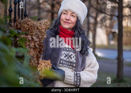Donna che gode di una soleggiata giornata invernale all'aperto, vestita con un comodo maglione e un cappello di pelliccia, circondata dalla natura, che irradia calore e felicità durante la stagione Foto Stock