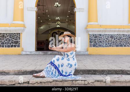 Una donna, che indossa un cappello e un vestito bianco e blu, si siede sui gradini di una chiesa e fa un selfie. Foto Stock
