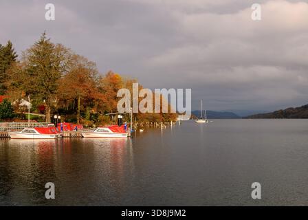 Scenario autunnale sul lago con barche ormeggiate alla luce del sole mattutino e alberi che diventano marrone dorato sotto un cielo nuvoloso Foto Stock