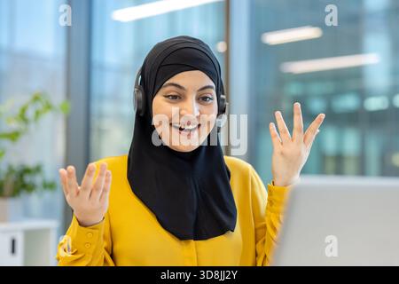 Donna musulmana che indossa un hijab nero e cuffie, sorridente e gestante mentre partecipa a una videochiamata online dal suo ufficio moderno, comunica e collabora virtualmente Foto Stock