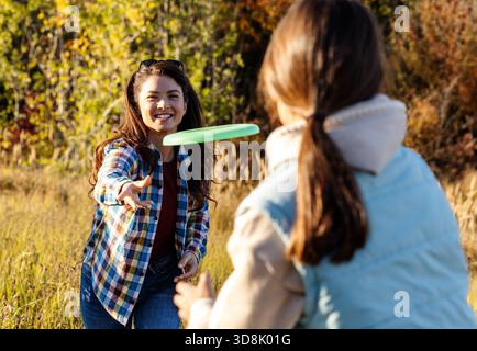 Mamma e figlia si divertono a giocare a frisbee nel campo soleggiato, circondati da erba alta e alberi. La luce del pomeriggio in autunno crea un'atmosfera calda Foto Stock