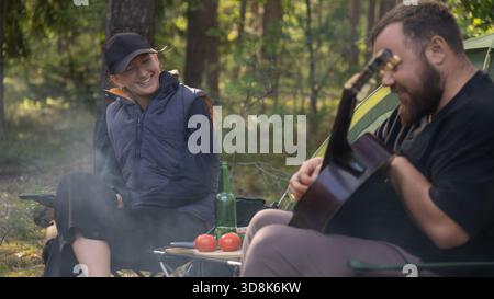 Coppia felice che si goda una giornata estiva in campeggio nella foresta, suona la chitarra vicino alla loro tenda, si rilassa mentre si fa un picnic e si gusta del cibo delizioso Foto Stock