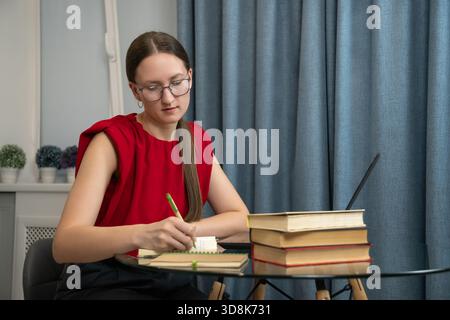 Una studentessa concentrata prende appunti sul notebook mentre impara a utilizzare il laptop e i libri in ufficio, si prepara all'esame o si esegue un compito Foto Stock
