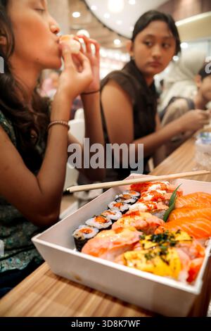 Un piatto di delizioso sushi di salmone giapponese assortito servito come menu per il pranzo preferito dalla famiglia presso un'area ristoro del supermercato con adolescenti seduti sullo sfondo Foto Stock