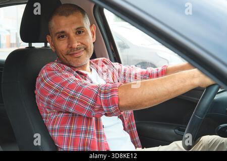 Uomo ispanico sorridente mentre guida un'auto, indossa una camicia rossa a quadri Foto Stock
