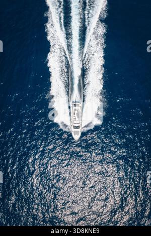 Vista aerea dall'alto verso il basso di uno yacht di lusso che naviga a velocità sostenuta sull'oceano scintillante e lascia una scia di bolle e onde alle spalle Foto Stock