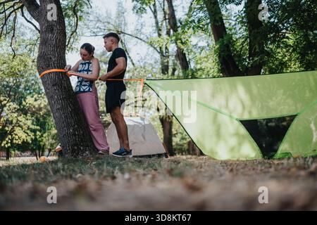 Coppia in campeggio all'aperto legando corda ad albero vicino a una grande tenda verde in una foresta Foto Stock