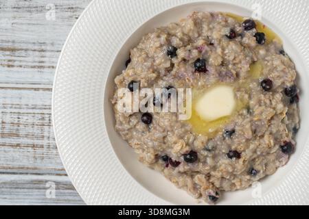 Porridge di farina d'avena semplice con mirtilli, burro e miele in piatto bianco su tavola di legno, primo piano, vista dall'alto. Il concetto di una colazione sana e p Foto Stock