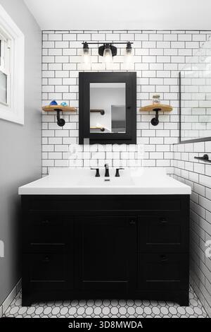 Bagno con armadietto nero su pavimento piastrellato bianco, lavandino nero su un piano di appoggio bianco e ripiani montati su una parete di piastrelle della metropolitana. Foto Stock
