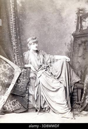 Ritratto artistico di una figura seduta in abiti greco-romani, posa su tappeti drappeggiati e frammenti architettonici. La composizione teatrale e lo stile classico suggeriscono spettacoli tardo vittoriani o edoardiani o rappresentazioni allegoriche. Da un album fotografico del tardo vittoriano a quello del primo edoardiano, che presentava molte carte di albume e carte de visites, fornendo una finestra sugli stili e le mode dell'epoca. Foto Stock