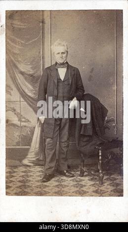 Un ritratto in bianco e nero di un uomo anziano in abito formale del XIX secolo, accanto a un tavolino con un cappotto drappeggiato. Indossa un cappotto lungo, un gilet e un cravatta. Lo sfondo presenta una tenda dipinta e vista panoramica; il pavimento e' piastrellato. Da un album fotografico del tardo vittoriano a quello del primo edoardiano, che presentava molte carte di albume e carte de visites, fornendo una finestra sugli stili e le mode dell'epoca. Foto Stock
