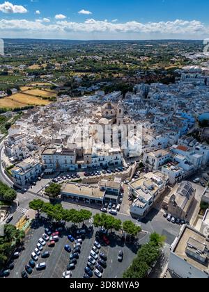 Veduta aerea degli edifici bianchi baciati dal sole raggruppati intorno alla chiesa torreggiante, in contrasto con i campi dorati e il vivace cielo blu, Locorotondo Foto Stock