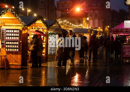 Le persone curiosano tra le bancarelle di un mercatino di Natale sotto la pioggia, con luci festose che illuminano la scena e riflessi sul terreno bagnato. Foto Stock