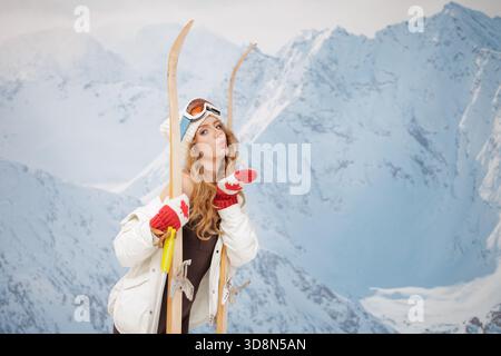 Giovane sciatore giocoso con l'abito invernale che soffia bacio con gli sci retrò sulle montagne innevate, perfetto per striscioni di vacanza sulla neve e concetti di saluto natalizio Foto Stock