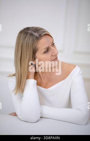 Donna caucasica di mezza età con lunghi capelli biondi, che indossa un top bianco, che guarda attentamente di lato, accettazione dei primi cambiamenti legati all'età Foto Stock