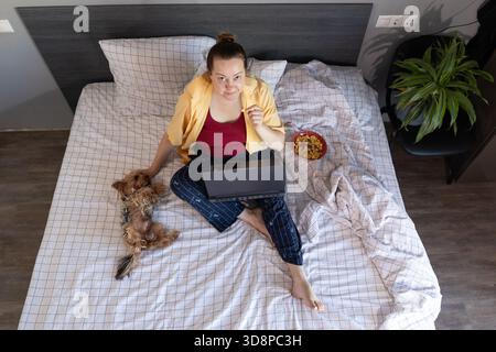 Una donna siede in un comodo letto con un computer portatile, gustando degli spuntini mentre il suo piccolo cane è accanto a lei. È rilassata e lavora da casa in un ambiente informale. Foto Stock