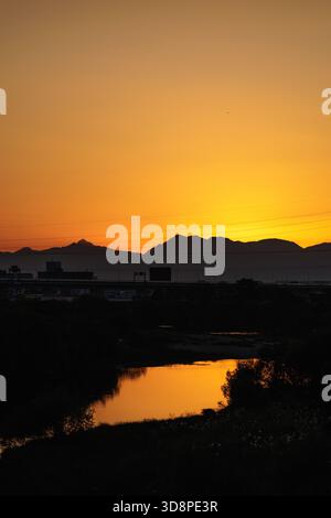As the sun dips behind mountains, a warm glow bathes city skyline in silhouette which encircles a quiet river mirroring sky's golden orange. Foto Stock