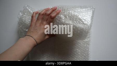 La mano femminile srotola un grande foglio di carta a bolle su sfondo bianco. Concetto di imballaggio di articoli fragili, protezione per la spedizione e consegna di g. Foto Stock
