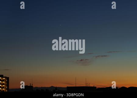 Venere splende luminoso sopra le silhouette di edifici che si trovano all'orizzonte del tramonto. I colori sfumati dell'arancione profondo trasversalmente al blu scuro su un cielo senza nuvole. Foto Stock