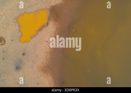 Scatto astratto di un drone che cattura una piscina tossica gialla vibrante su un letto di lago essiccato. Il contrasto tra terra incrinata e acqua inquinata torbida illustra la gravità della crisi ecologica. Foto Stock