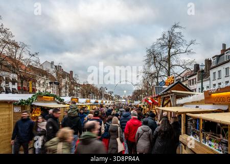 Bruxelles, Belgio - 30 novembre 2025: Folle che camminano attraverso il mercatino di Natale fiancheggiato da bancarelle di legno, con la ruota panoramica delle meraviglie d'Inverno nella ba Foto Stock