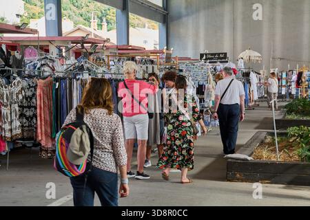 Dubrovnik. Croazia - 2 dicembre 2025: Diversi turisti, tra cui una donna con uno zaino arcobaleno, curiosano in un mercato coperto per i vestiti estivi e così via Foto Stock