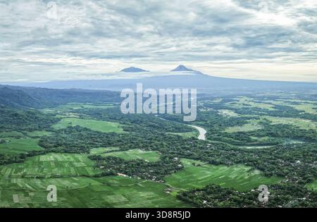La vista aerea dei campi di riso color smeraldo incontra l'orizzonte, dove il maestoso Monte Merapi e il Monte Merbabu sono sorvegliati sotto un vasto cielo nuvoloso, Yogyakarta Foto Stock