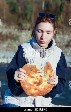Una giovane donna in abiti caldi tiene una focaccia tradizionale appena rotta con un buco mentre è seduta su una sponda rocciosa in autunno Foto Stock