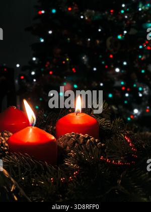 Primo piano di una corona dell'avvento con due candele rosse accese e luci sfocate sull'albero di Natale Foto Stock