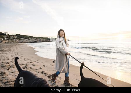Donna sorridente mentre cammina i suoi due cani neri al guinzaglio lungo una spiaggia sabbiosa Foto Stock