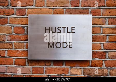 Su una parete in mattoni rustici è montato un robusto cartello in metallo che mostra la scritta Hustle mode. Questa impostazione irradia motivazione ed energia, incoraggiando il profarmaco Foto Stock
