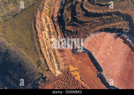 Colpo astratto di un drone di una miniera a cielo aperto. Le terrazze di argilla rossa, i pali di roccia bianca e un piccolo camion mettono in risalto l'enorme scala dell'estrazione industriale e dello sviluppo delle materie prime. Foto Stock