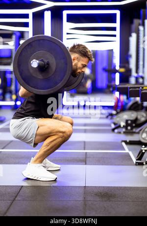 Un uomo esegue uno squat con un pesante barbell in una palestra elegante e vivace, mostrando dedizione e forza durante un allenamento. Foto Stock