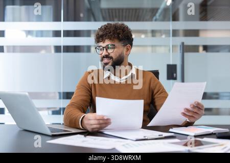 Giovane uomo d'affari indiano seduto alla scrivania dell'ufficio, sorridente mentre esamina documenti e lavora su un laptop, gestendo attività in un ambiente aziendale professionale Foto Stock
