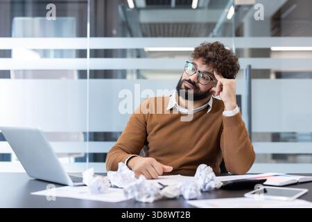 Uomo frustrato con barba e bicchieri che si sente stressato e sperimenta il blocco dello scrittore in una moderna scrivania, circondato da carta sgualcita, cercando di trovare idee nuove Foto Stock