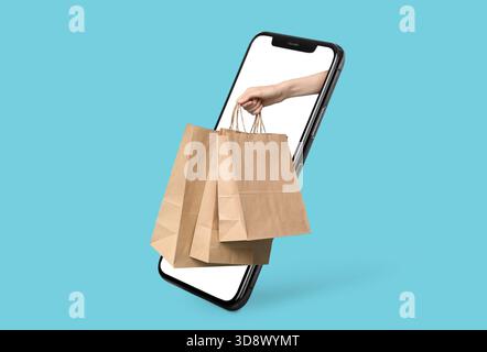 Mano con i sacchetti di carta che escono dallo schermo dello smartphone su sfondo blu Foto Stock