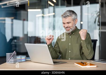 Uomo d'affari maturo che ha ottenuto risultati e successi, tifa con pugni serrati e guarda i risultati positivi su un computer portatile in un moderno ambiente di lavoro Foto Stock