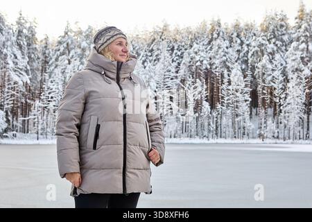 Una donna si trova vicino al lago ghiacciato, indossa un cappotto invernale e un cappello a maglia, circondato da alberi innevati e gode di un panorama invernale. Foto Stock