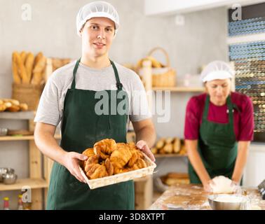 Il giovane panettiere in uniforme si trova in una piccola panetteria privata che offre croissant caldi Foto Stock