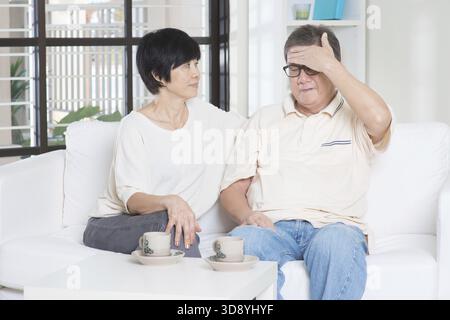 Ritratto di un uomo asiatico maturo che ha mal di testa, seduto sul divano con la moglie a casa, pensionato anziano che vive in casa Foto Stock
