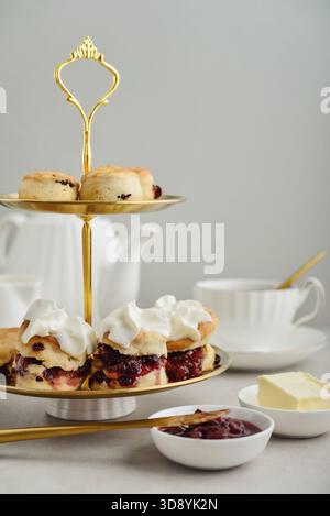 Stand dorato a due piani con scones fatti in casa serviti con burro e marmellata e tazze da tè in porcellana bianca sullo sfondo, primo piano Foto Stock