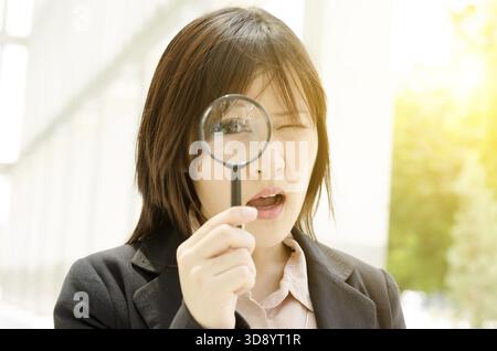 Giovane donna d'affari asiatica che guarda attraverso la lente d'ingrandimento, in un ambiente d'ufficio, luce naturale dorata del sole sullo sfondo Foto Stock