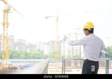 Ritratto di un ingegnere appaltatore indiano asiatico sorridente con elmetto che indica il cantiere Foto Stock