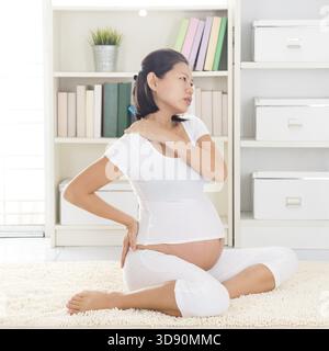 Dolore alla spalla o al collo. Donna asiatica incinta di otto mesi che le tiene la schiena e la spalla seduto sul pavimento di casa Foto Stock