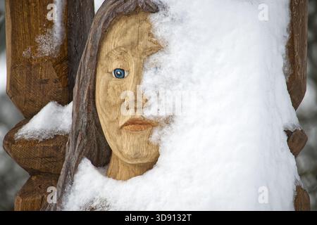 Una statua scolpita in legno di un volto umano si abbina da sotto un velo di neve. L'occhio blu si distingue, creando una tranquilla e misteriosa scena invernale con il natur Foto Stock