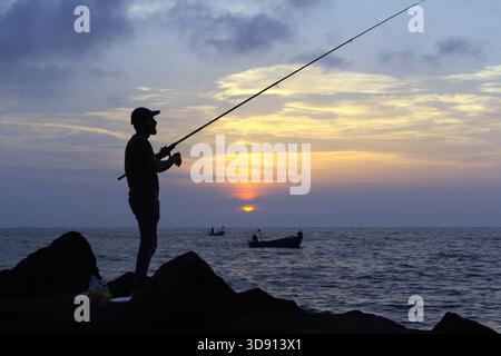 • Silhouette di Fisherman focalizzata contro un tramonto morbido Un pescatore solitario in silhouette si erge sulle rocce, gettando la sua linea nel mare. Foto Stock
