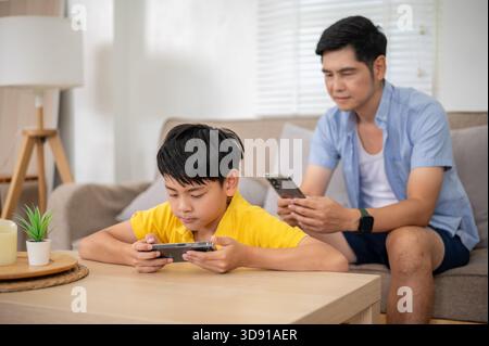 Il padre asiatico e il figlio della scuola elementare guardano video sullo smartphone mentre sono seduti nel soggiorno della loro casa. Attività online, navigazione in Internet Foto Stock