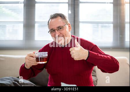 Vecchio bianco caucasico che indossa occhiali che tiene il caffè e prepara il pollice seduto sul divano in soggiorno. Versare o far gocciolare le mani. Foto Stock