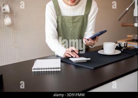 Primo piano di una barista o di una proprietaria in grembiule verde che tiene in mano lo smartphone mentre utilizza una calcolatrice sulla barra del bancone. Caffetteria o caffetteria, piccola impresa. Foto Stock