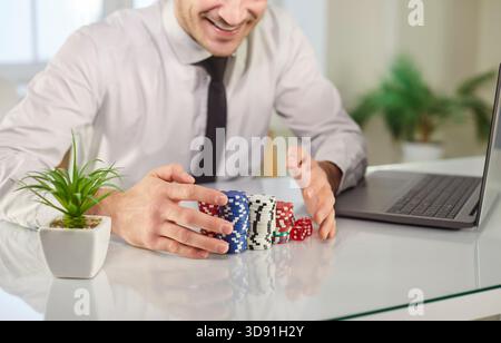 Uomo d'affari entusiasta che celebra la vittoria del poker online, impilando patatine colorate e dadi alla scrivania. Foto Stock
